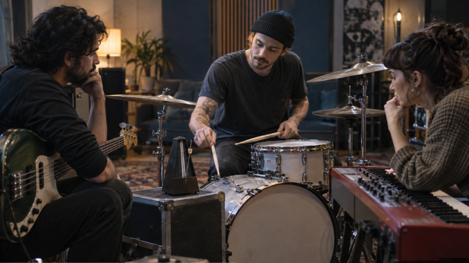 A drummer, bassist, and keyboardist listen around a metronome while deciding how the song should move.