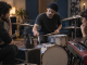 A drummer, bassist, and keyboardist listen around a metronome while deciding how the song should move.