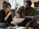 A songwriter and session guitarist discuss a marked song arrangement beside microphones in a recording room.