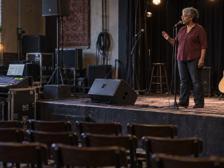 A vocalist listens and gestures to a sound engineer during a microphone check in an empty small venue.