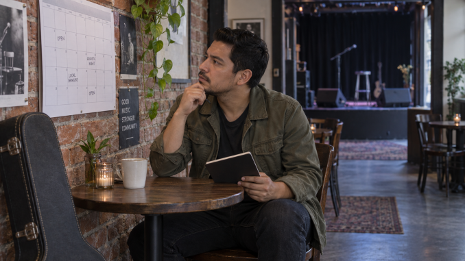 A guitarist studies a venue calendar beside the small stage while holding a notebook.