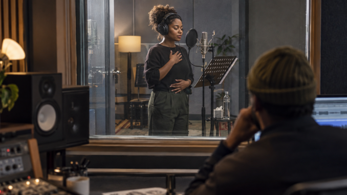 A vocalist checks her breathing beside a prepared studio microphone while the producer waits in the control room.