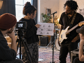 An independent artist and small video crew arrange a performance shot using a storyboard in a modest rehearsal space.