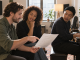 A songwriter reviews a clean sectioned lyric sheet with a singer and pianist during rehearsal.