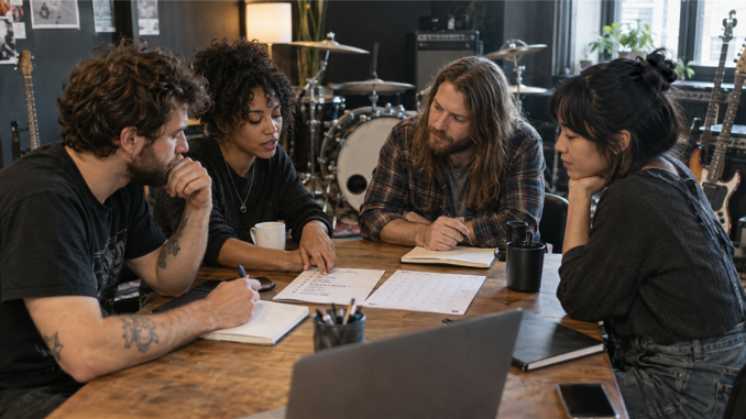 Four band members sit around a rehearsal-room table reviewing a concise decision list and shared calendar.